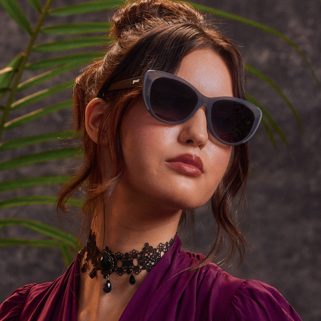 An elegantly dressed woman in translucent gray cat-eye sunglasses with black gradient lenses.
