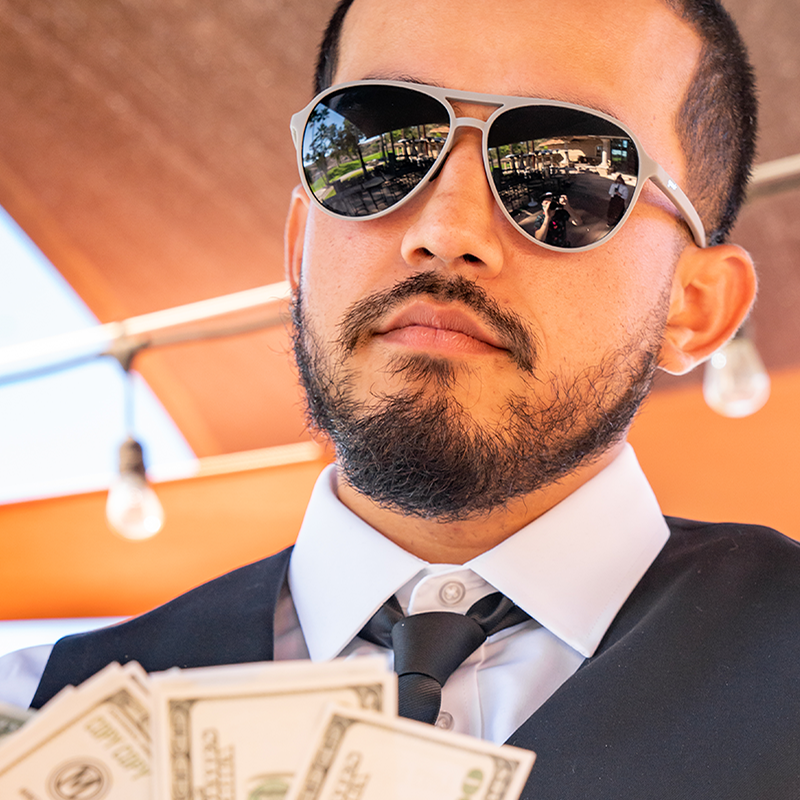 A man in a tie and waistcoat at a golf clubhouse wears gray aviator sunglasses with black lenses, fanning himself with cash.