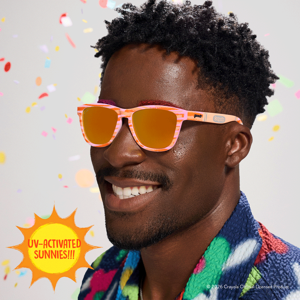 Young man smiles, wearing Crayola goodr UV-activated sunglasses with bone-to-magenta frames and amber reflective lenses.