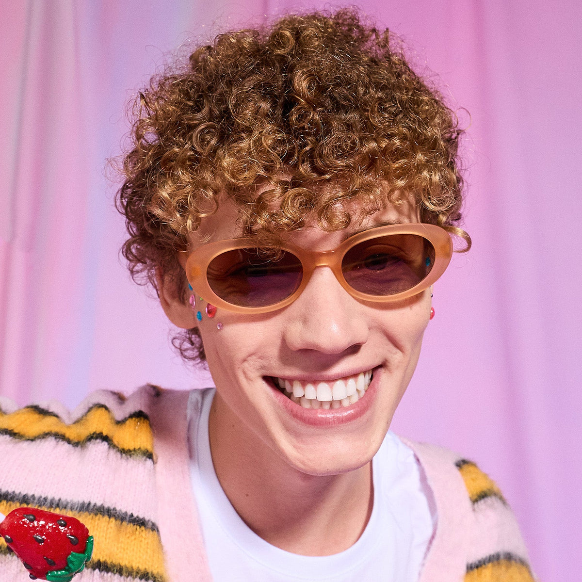 A young man with strawberry blond curls smiles confidently in a pair of translucent pale peach oval Beat G sunglasses.