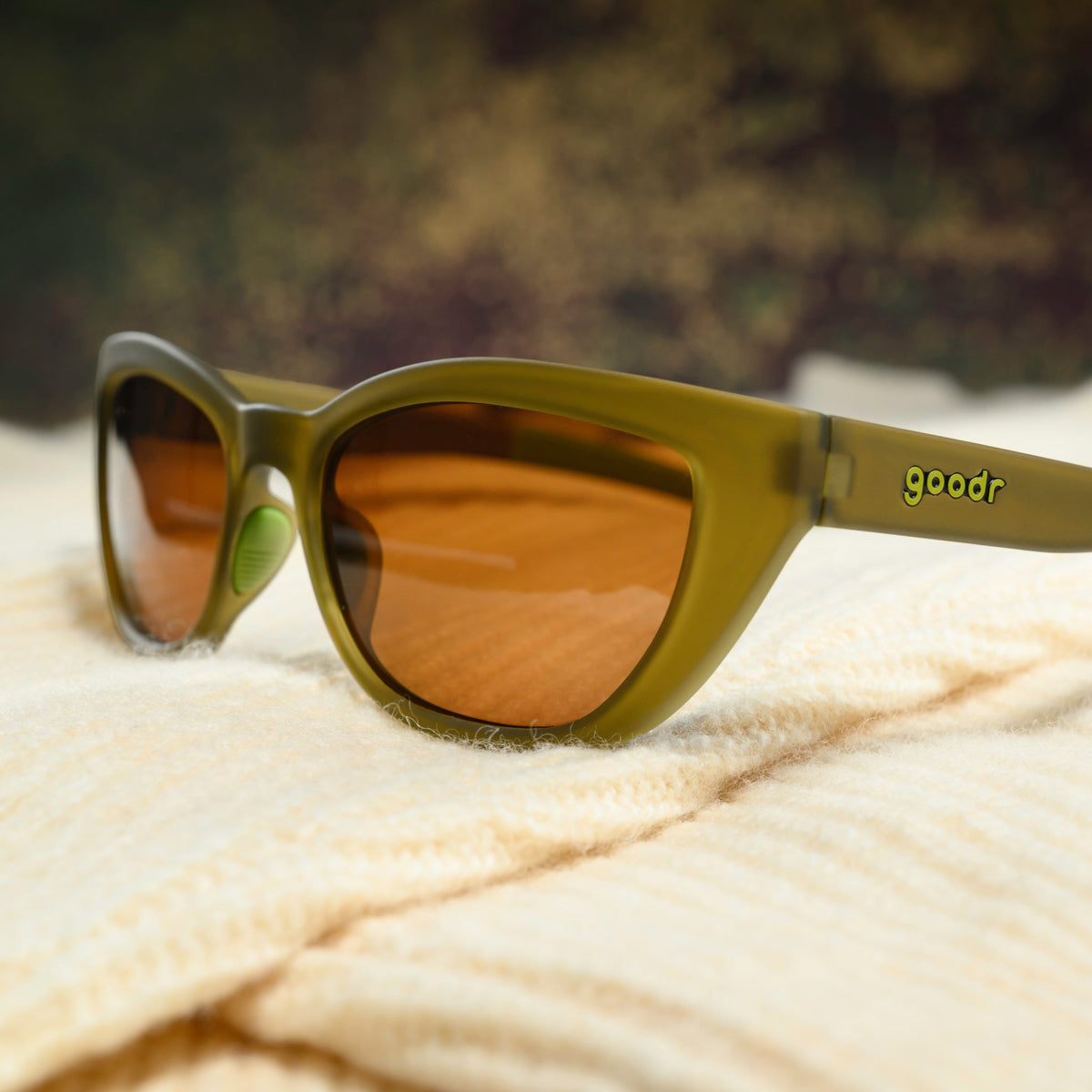 A pair of translucent olive-green cat-eye sunglasses with brown non-reflective lenses sits on a cream-colored sweater.