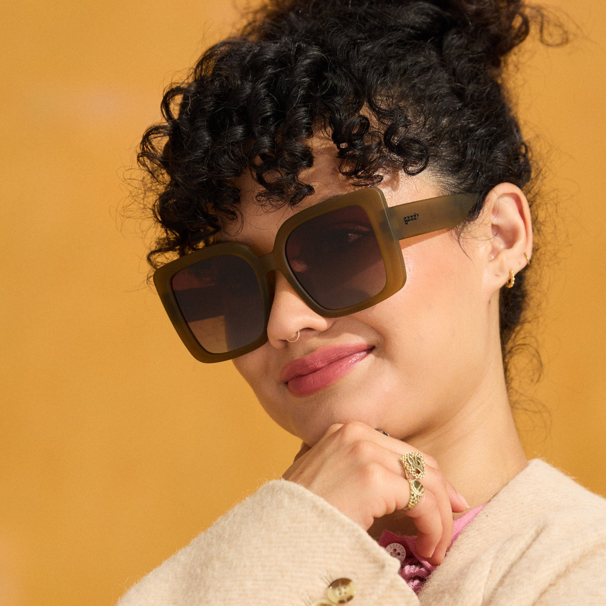 A woman with dark curls poses confidently in a pair of oversized square Amp G sunglasses in a translucent dusted-ochre hue.