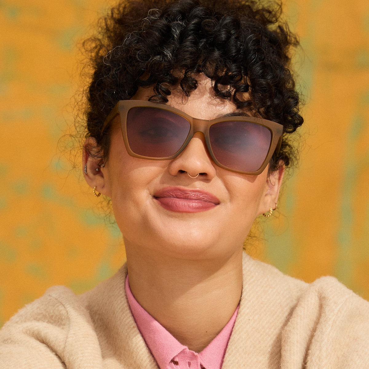 A woman with dark curls smiles warmly in a pair of ochre-yellow angular cat-eye sunnies against a golden ochre background.