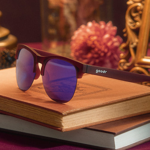 Three-quarter angle view of translucent purple half-rim sunglasses with purple reflective lenses on top of stacked books.