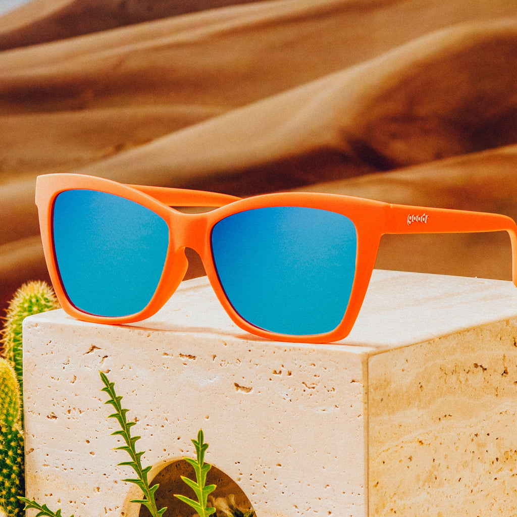 Three-quarter angle view of red angular sunglasses with blue reflective lenses on top of a clay block in front of a desert backdrop.