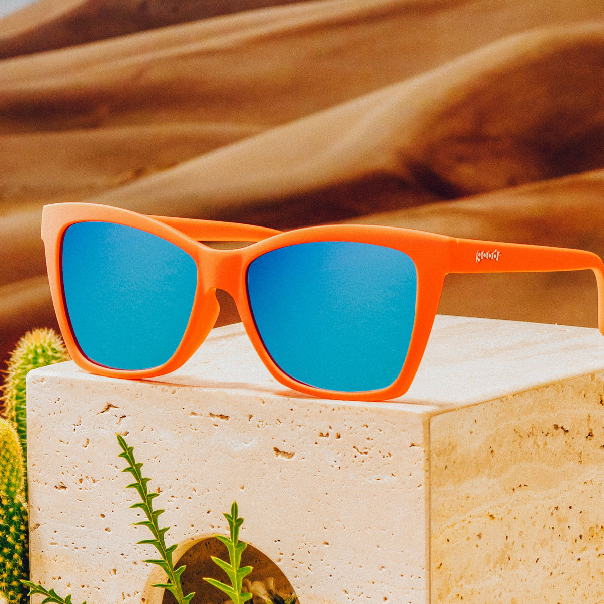 Three-quarter angle view of red angular sunglasses with blue reflective lenses on top of a clay block in front of a desert backdrop.