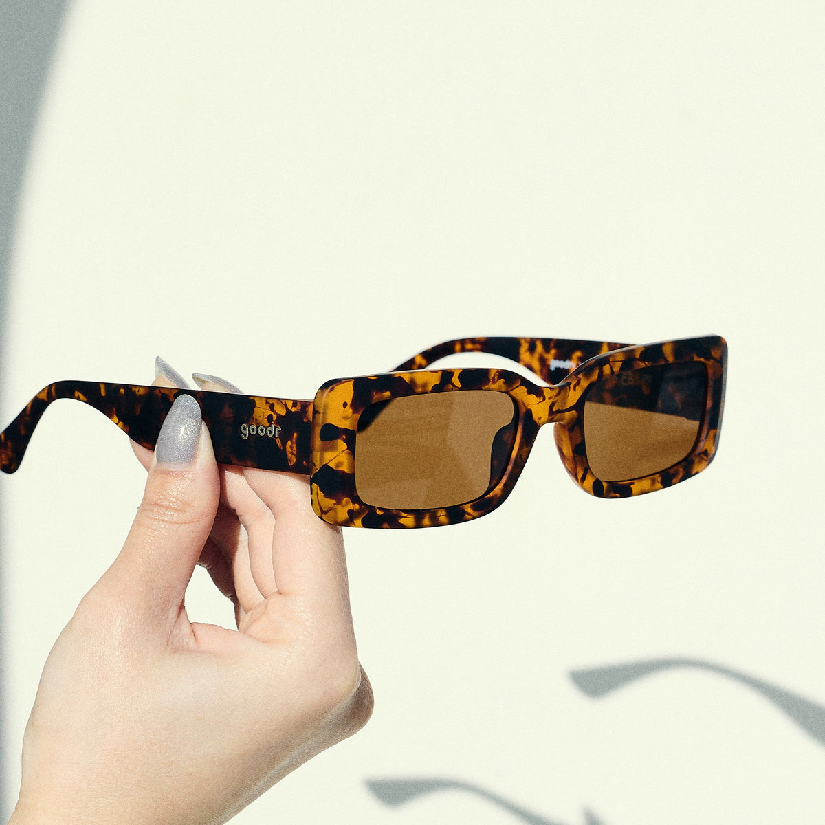 A manicured hand holds up a pair of rectangular sunglasses with brown polarized lenses in front of an off-white backdrop.