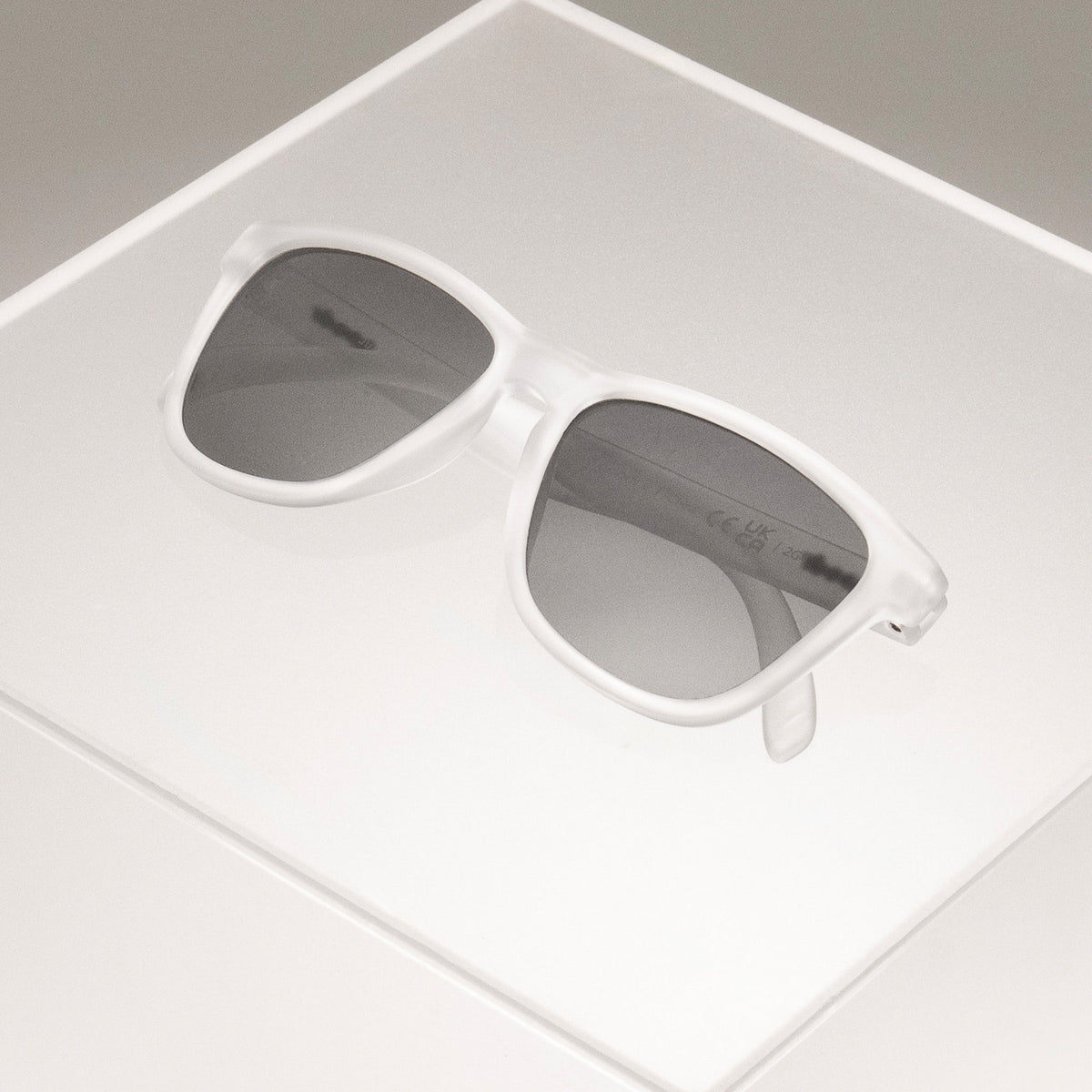 Clear sunglasses in a classic frame shape with black non-reflective lenses folded, sitting atop a clear plastic box.
