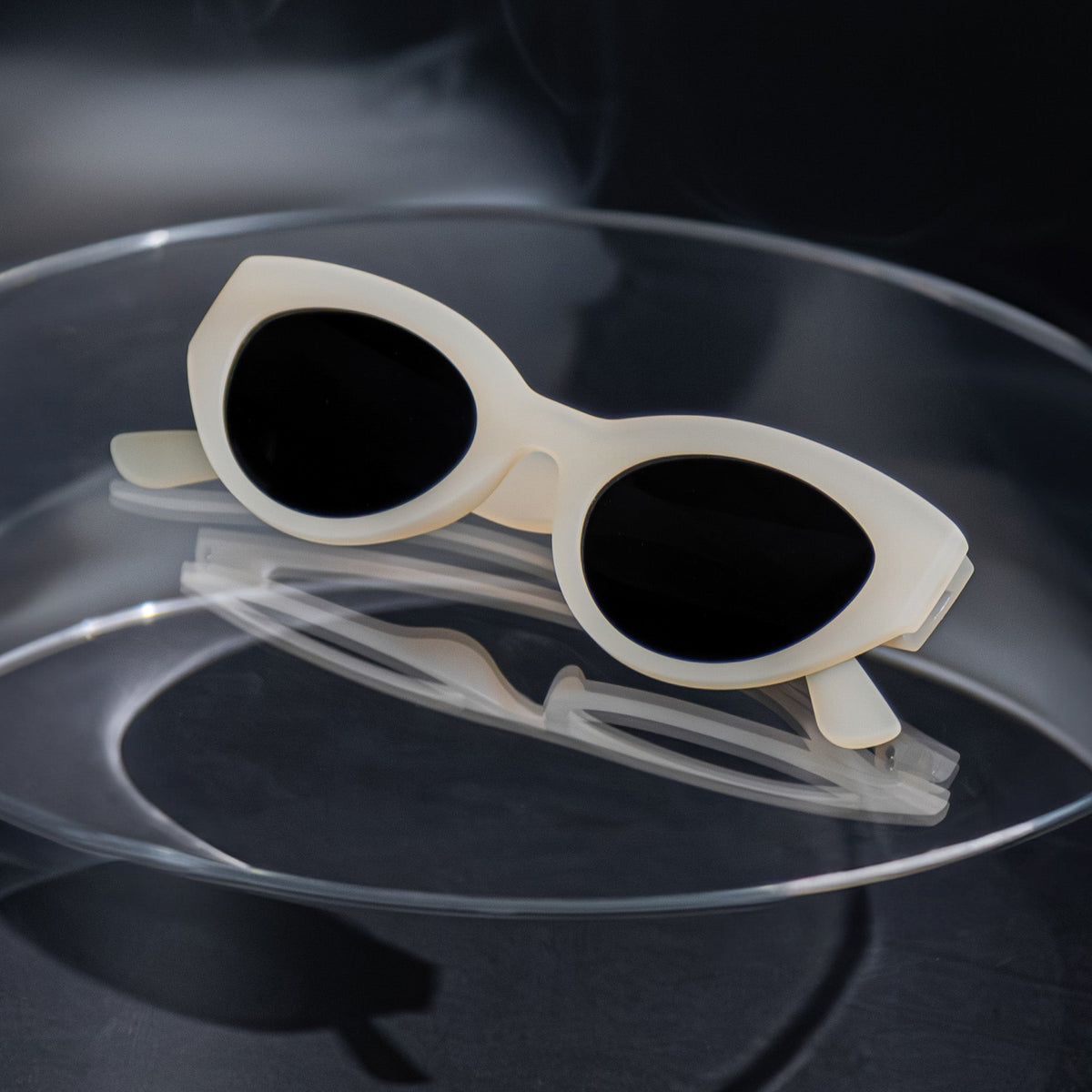 Close-up of translucent soft cream, retro-inspired angled cat-eye sunglasses with black non-reflective lenses, folded on a clear table.