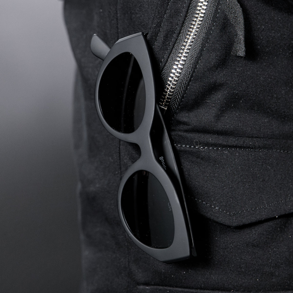 Close-up of translucent black, retro-inspired angled cat-eye sunglasses with non-reflective lenses hanging from a black zippered pant pocket.