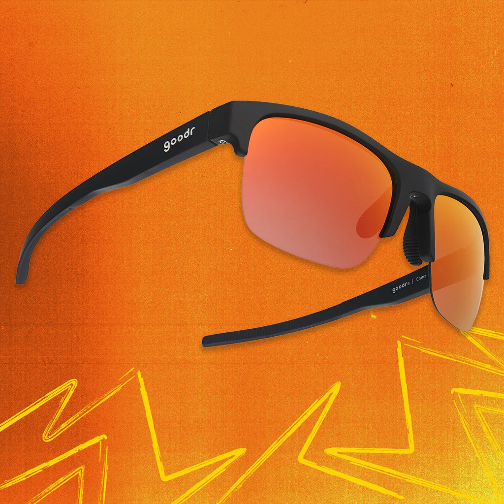 Three-quarter angle view of black shades w/ half-rim frames+orange reflective lenses tilted up on an orange background.