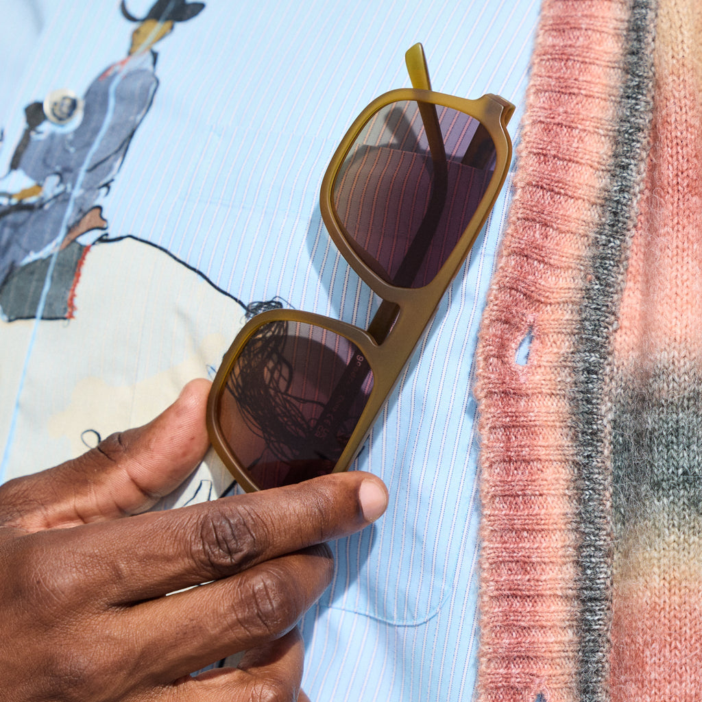 Close-up shot of a man’s hand holding a pair of translucent dusted-ochre Retro G sunglasses close to his chest pocket.