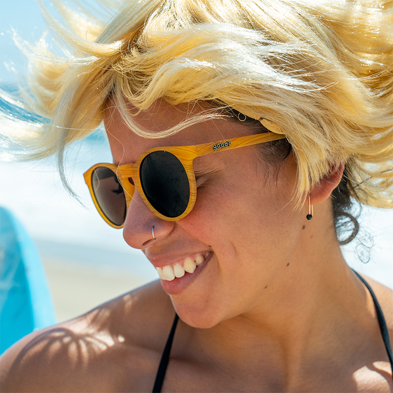 A woman in a short blonde wig wearing brown wood grain patterned circular sunglasses with brown lenses looks off, smiling.