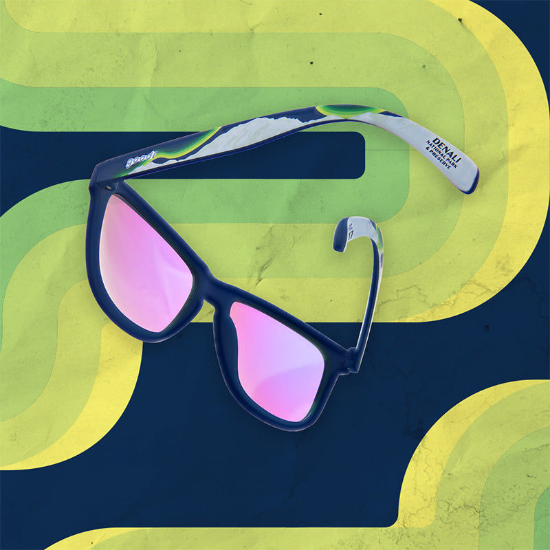 A pair of blue, green and yellow goodr sunglasses with magenta lenses on a blue and green background, with "Denali National Park & Preserve" on the end of the temple.
