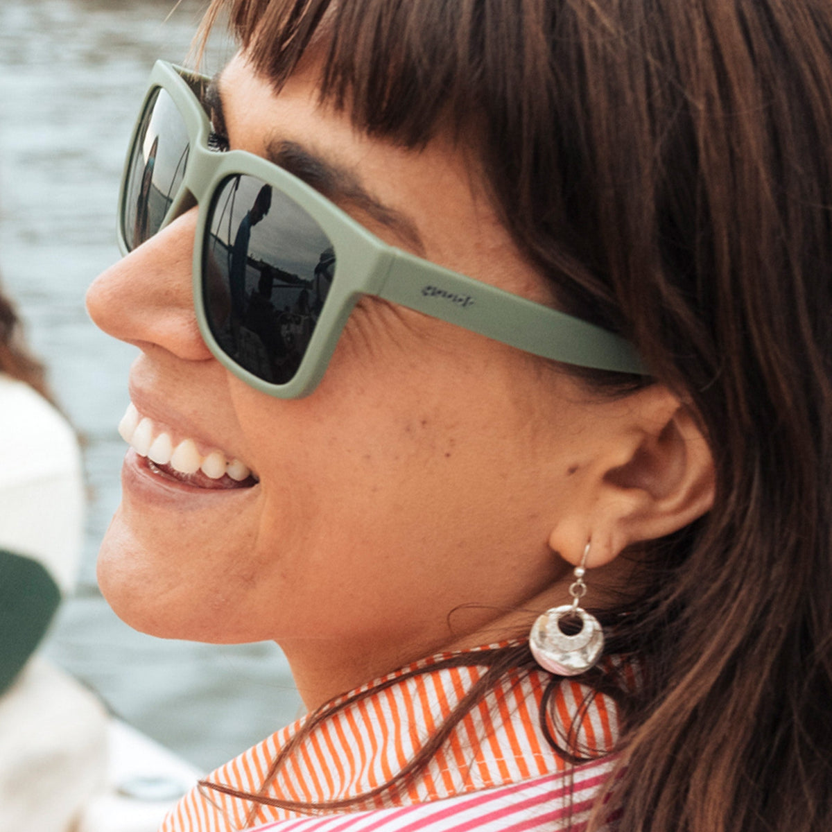 Three-quarter angle view of a laughing young woman with brown hair wearing dark green sunglasses with green lenses.