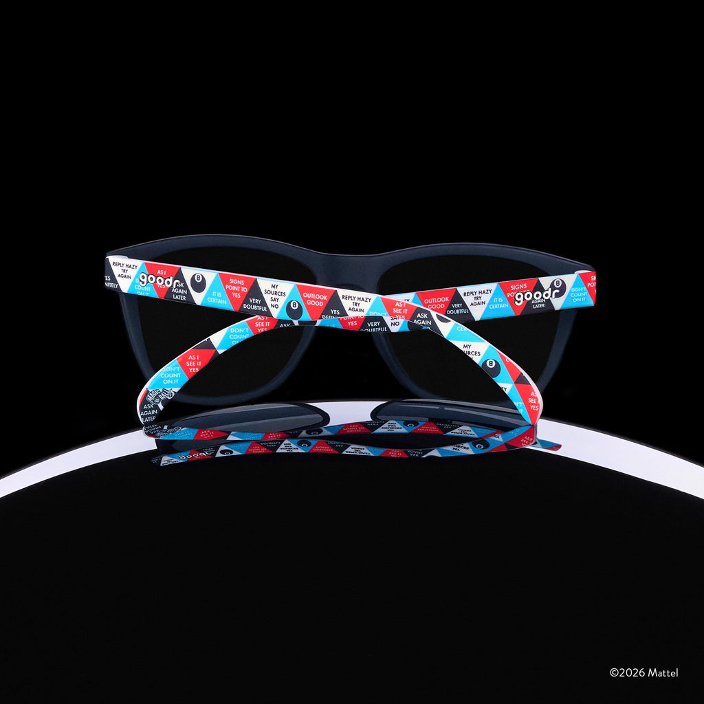 A pair of Magic 8 Ball-themed goodr sunglasses is perched atop the curve of an 8 ball, photographed from behind.