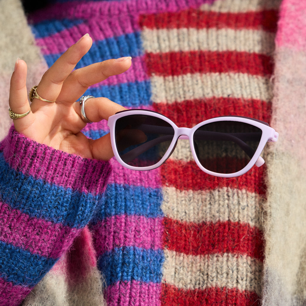A woman's hand holds a pair of lavender Pounce Gs out in front of her chest. She wears a colorful, fuzzy, striped sweater.