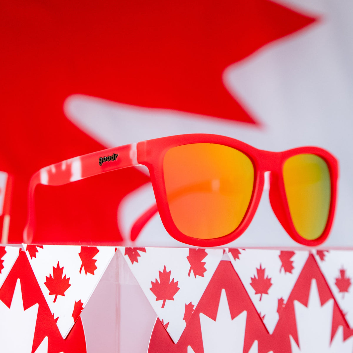 Angled view of red shades w/ a Canadian flag pattern+burnt orange reflective lenses on a podium w/ a maple leaf pattern.